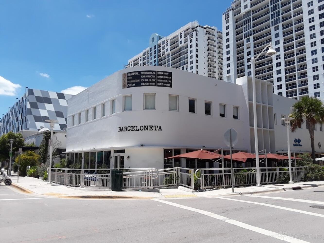 A restaurant with curved white exterior walls, a corner entrance, and outdoor seating under red umbrellas at Barceloneta Miami.