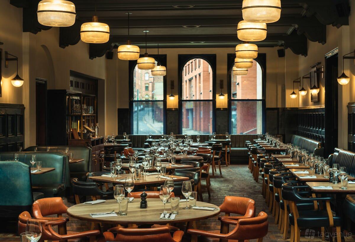 A restaurant dining room with leather chairs, set tables, large arched windows, and warm pendant lighting at Hawksmoor Manchester.