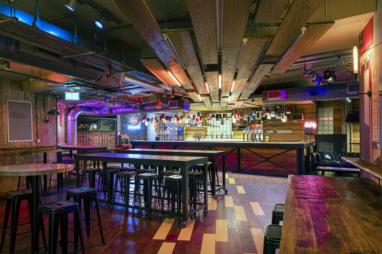 A bar with industrial lighting, high wooden tables, and colorful ceiling accents at Shuffleboard Bar at Generator London.