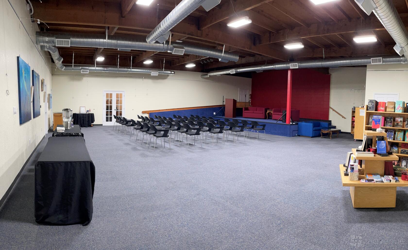 An event venue with rows of black chairs facing a raised stage, exposed ductwork, and a carpeted floor at Hen House Literacy Center.