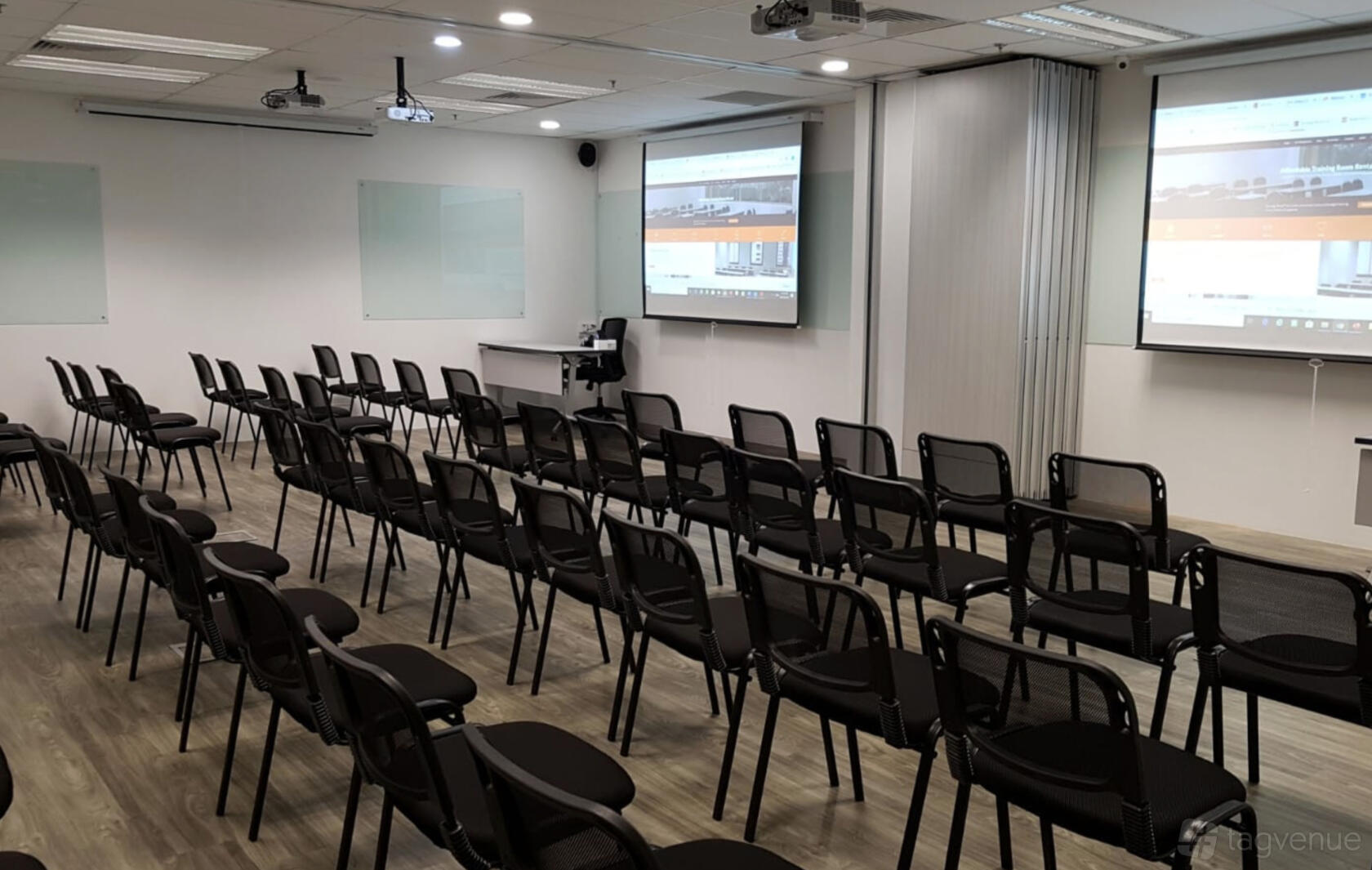 A training room with rows of black chairs, dual projection screens, and wood flooring at Synergy Room Havelock 2.