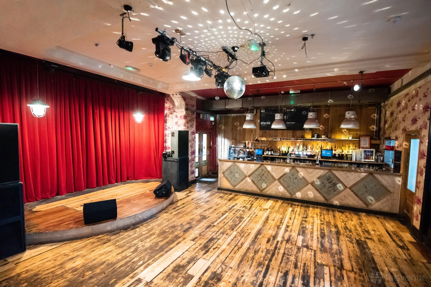 An event space in a nightclub with a wooden stage, disco ball, red curtains, and wooden floors at Deaf Institute.