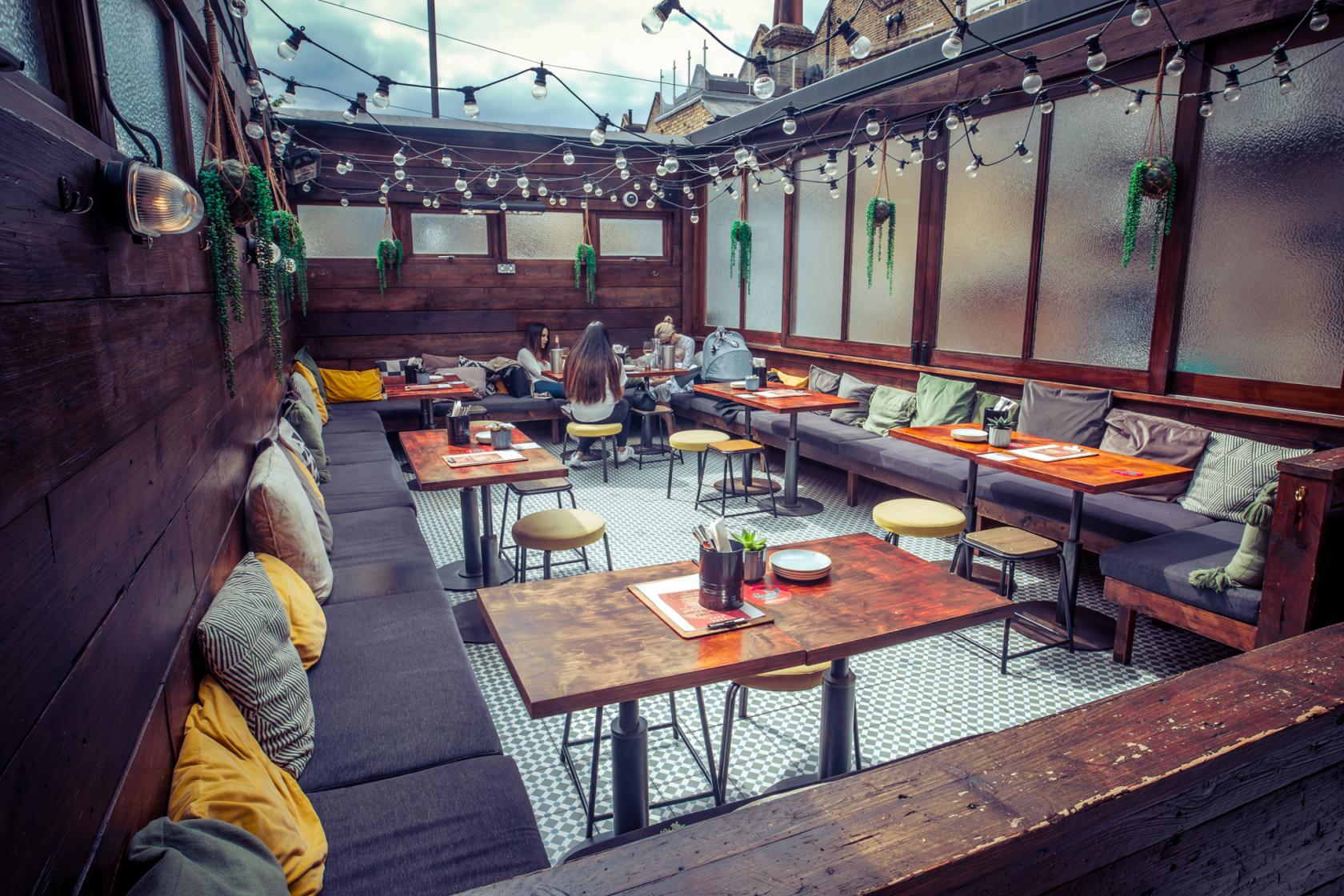A terrace bar with wooden walls, string lights, bench seating with cushions, and potted plants at Vagabond Northcote Road.