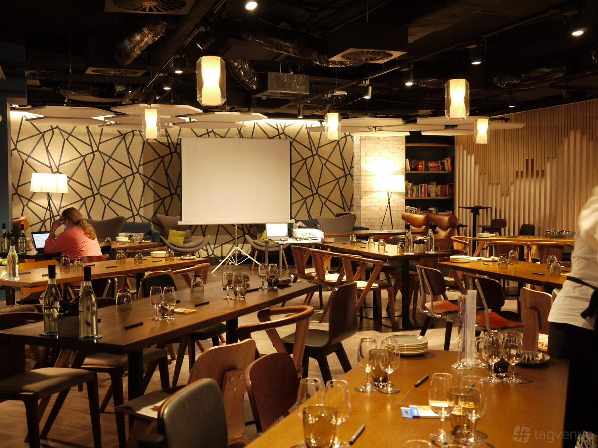 A conference centre lounge with wooden tables, patterned accent wall, ceiling pendant lights, and a projector at Cliftons Sydney Spring St.