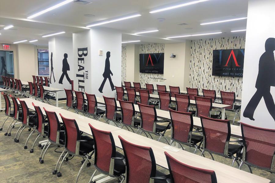 A meeting room with rows of red mesh chairs, long white tables, and Beatles-themed wall art at Midtown East Jay Suites.