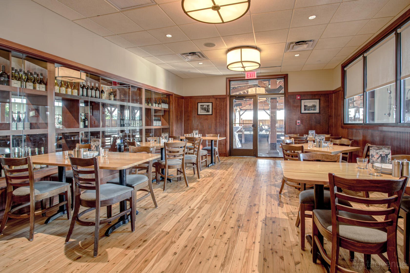 A dining room with wooden tables, matching chairs, large windows, and a display wall with wine bottles at Pinstripes - South Barrington.