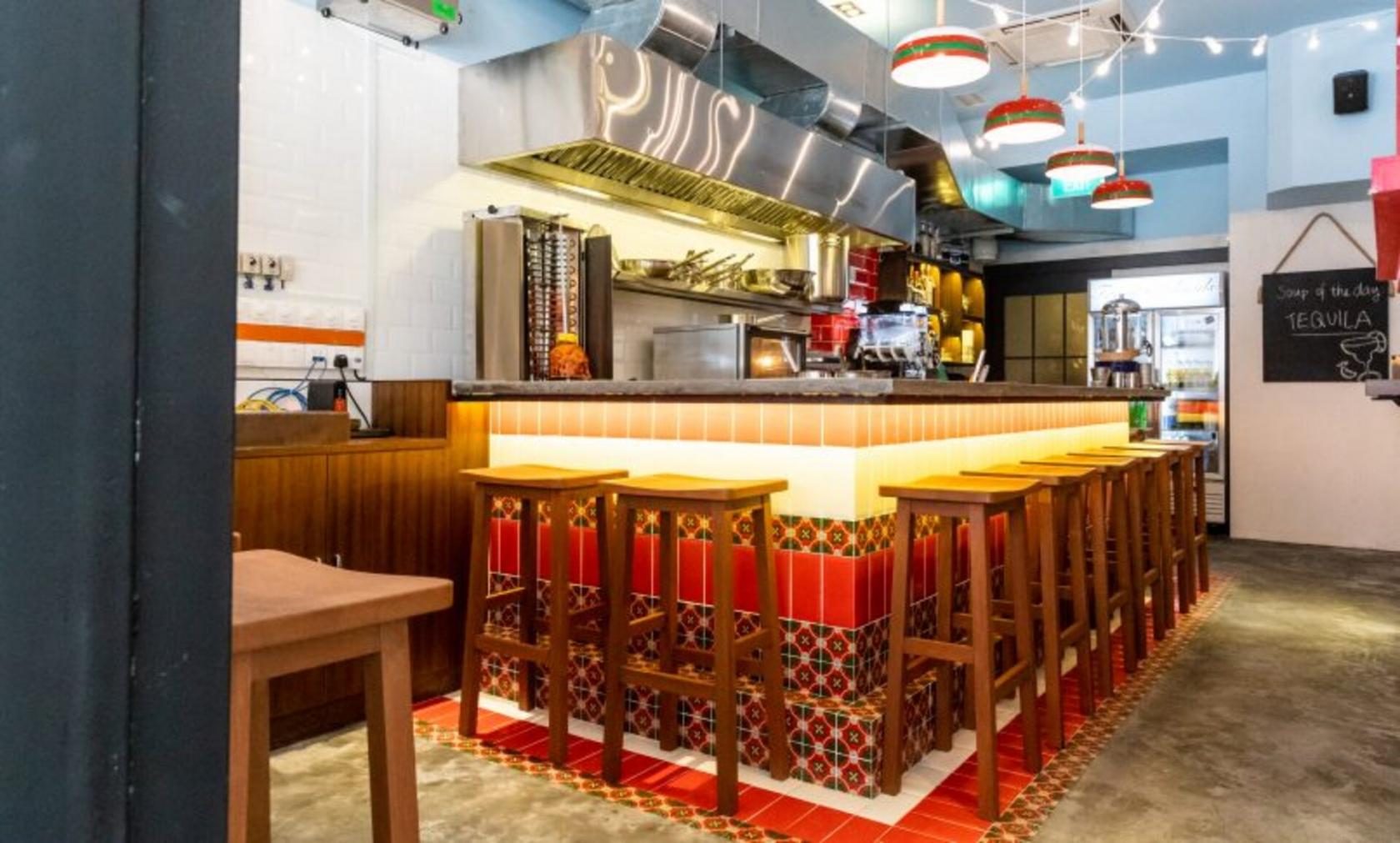 A brightly lit restaurant with a tiled bar counter, wooden stools, and open kitchen at Papi's Tacos.