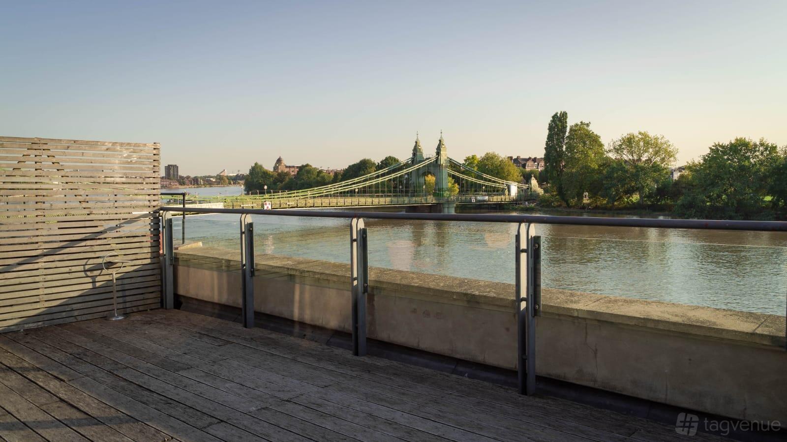 An event venue with a wooden deck and glass railing overlooking a river and suspension bridge at Auriol Kensington Rowing Club.