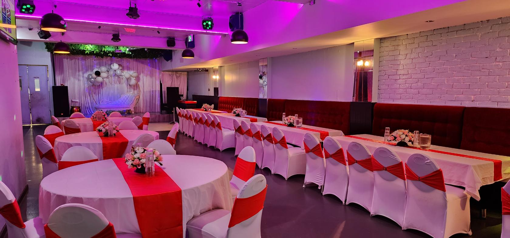 A function room with round tables and long banquet seating, white chair covers with red sashes, and pink uplighting at Third Eye Bar Rooftop Restaurant.
