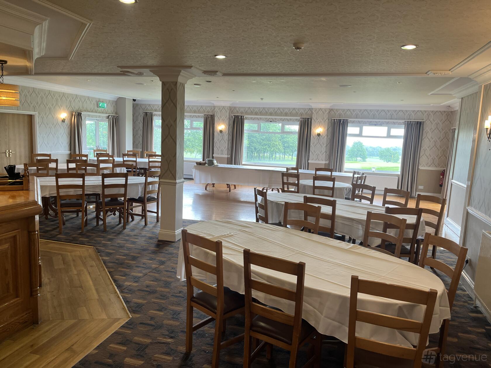 An event venue with large windows, round and rectangular tables with white linens, and wooden chairs at The Manor Golf Club.