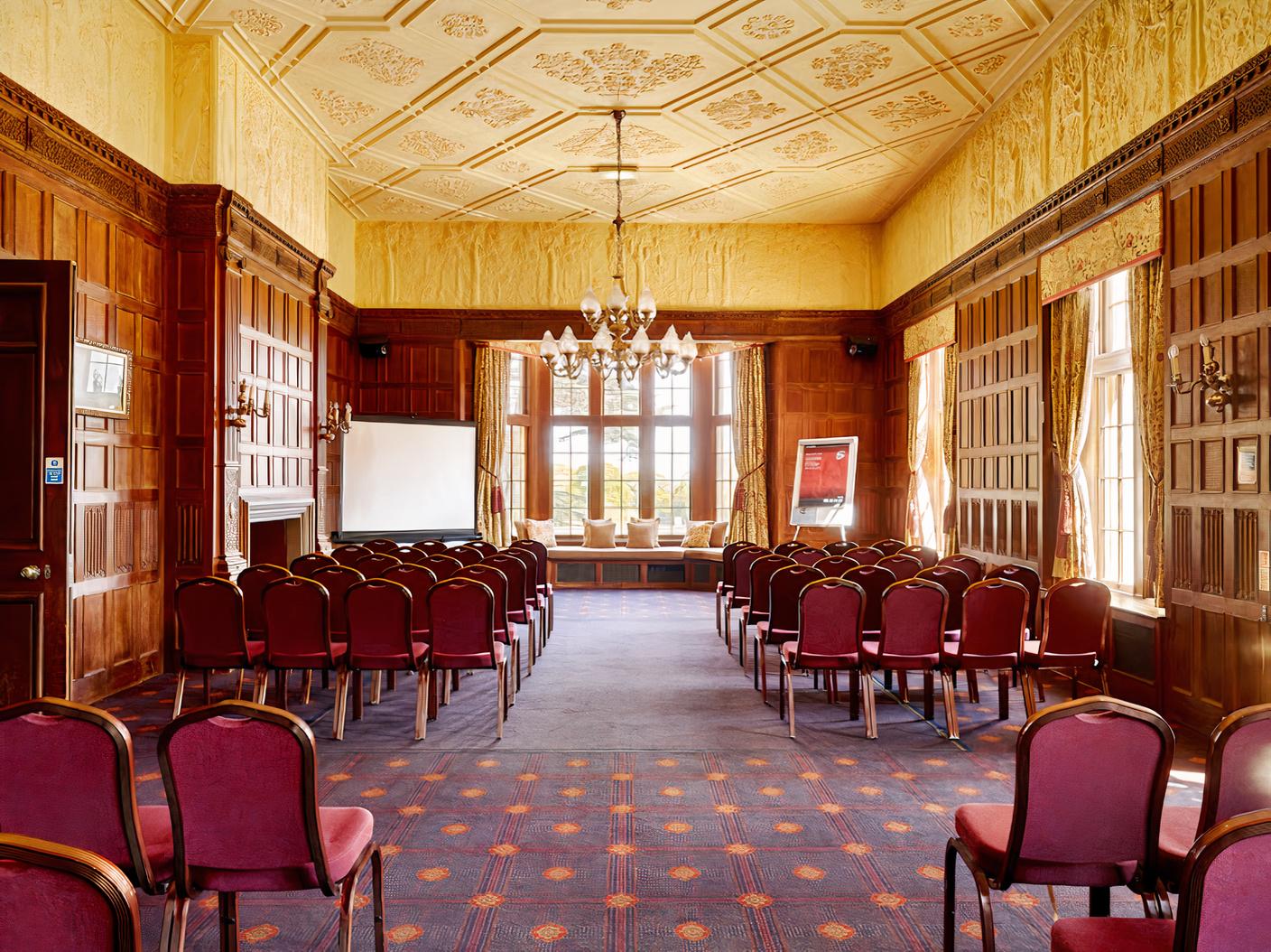 An event space with wood-paneled walls, chandeliers, red cushioned chairs, and tall windows at Dumbleton Hall Hotel.