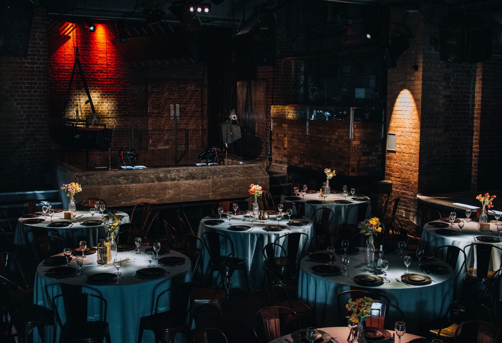An event venue with exposed brick walls, dramatic lighting, and round tables set with floral centerpieces at fabric industrial arts events venue.
