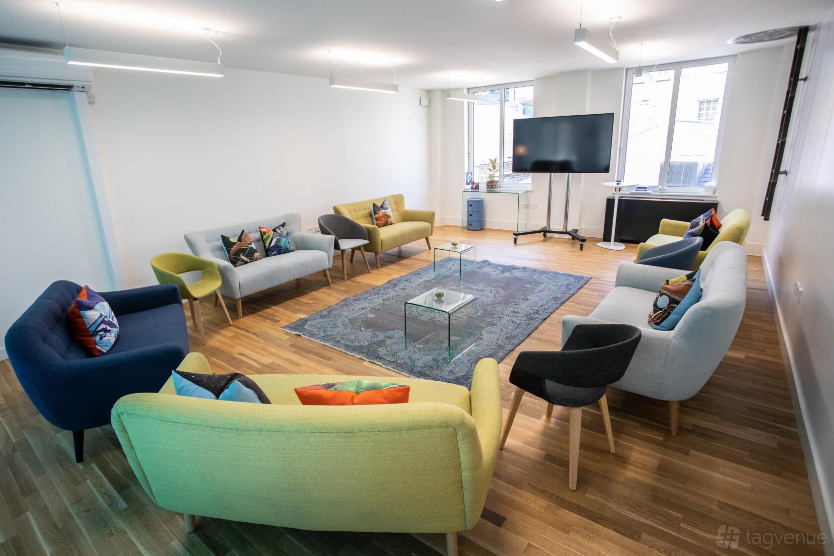 A meeting room with colorful sofas arranged in a circle, large windows, and a TV screen at Wallacespace Spitalfields.
