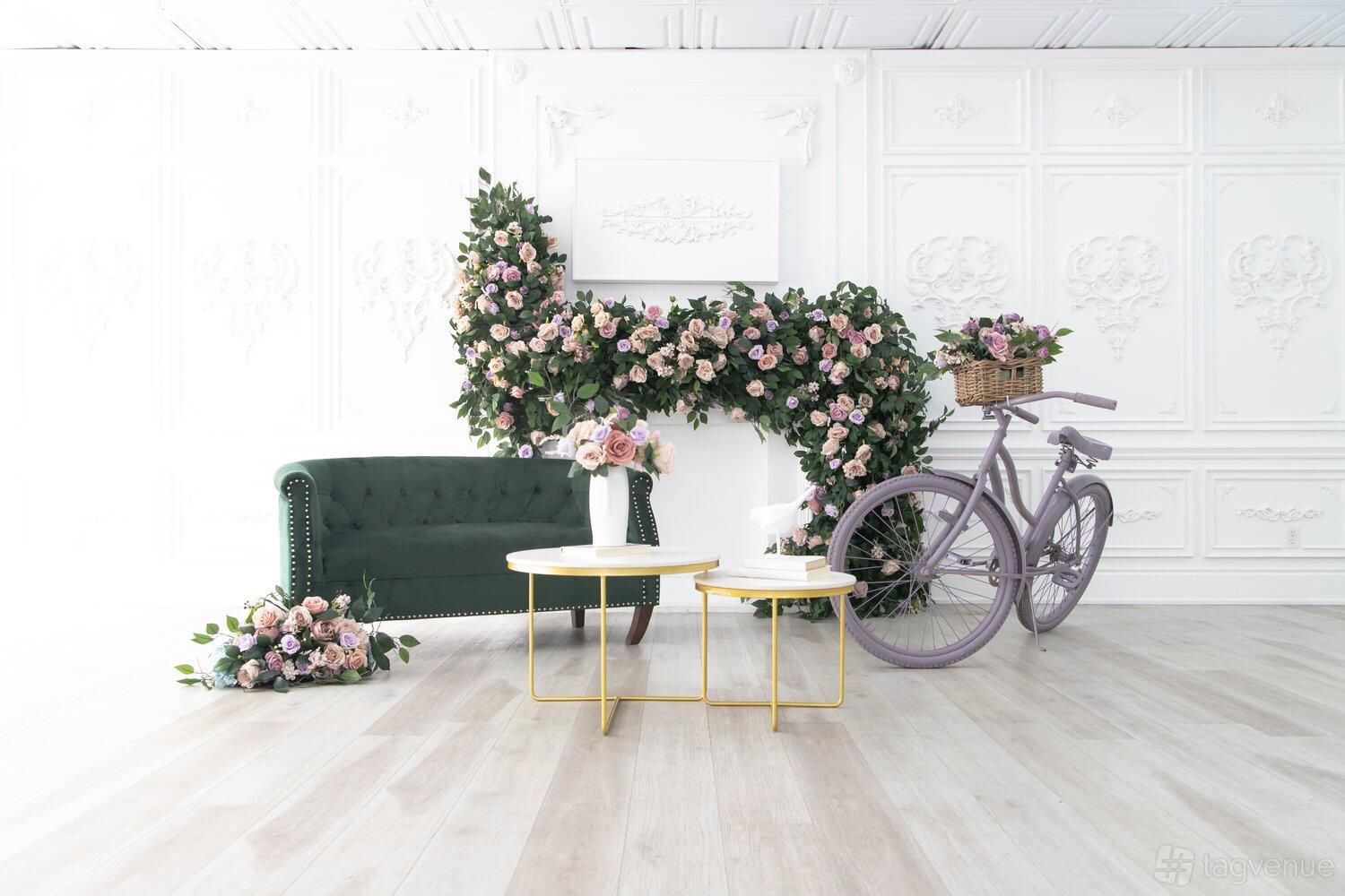 A photo studio with white ornate walls, pale wood floors, floral arrangements, a green velvet sofa, and a lilac bicycle at Vision Room Studios.