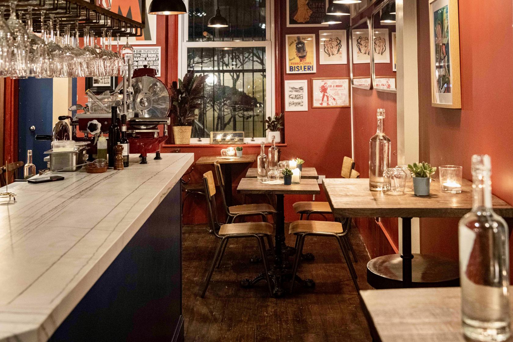 A cozy bar with a marble countertop, hanging glassware, wooden tables, and candlelit seating at Il Maremmano.