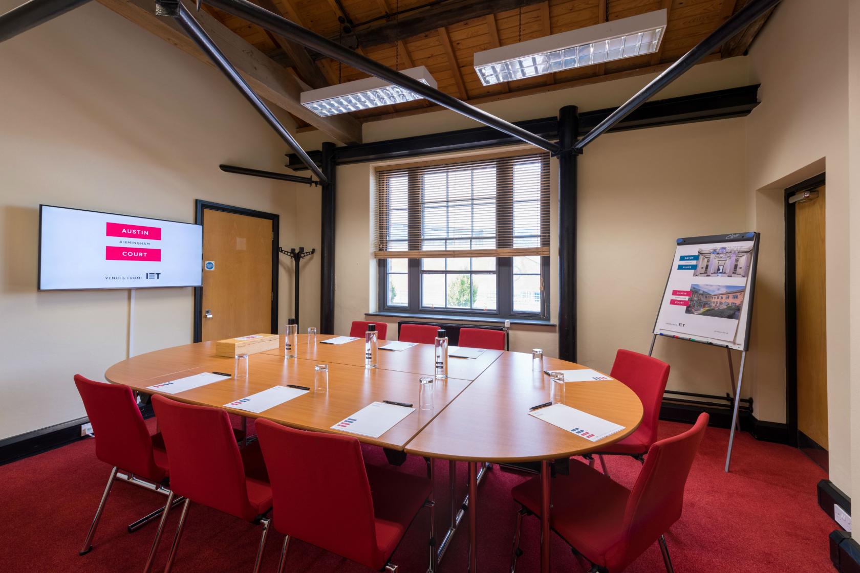 A conference centre room with a round table, red chairs, large windows, and a presentation screen at IET Birmingham: Austin Court.