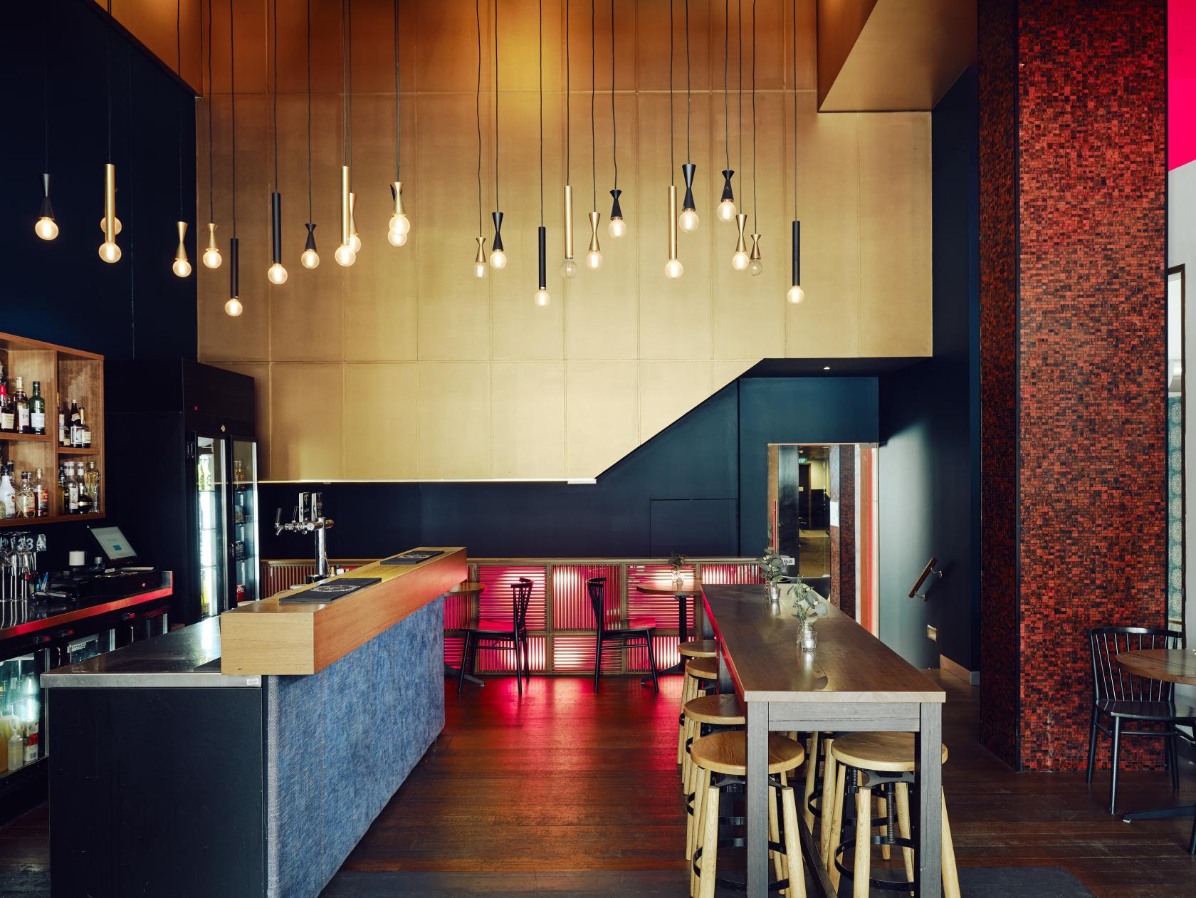 A hotel bar with exposed bulb pendant lights, a wood bar counter, and stools at Saint Kilda Beach Hotel