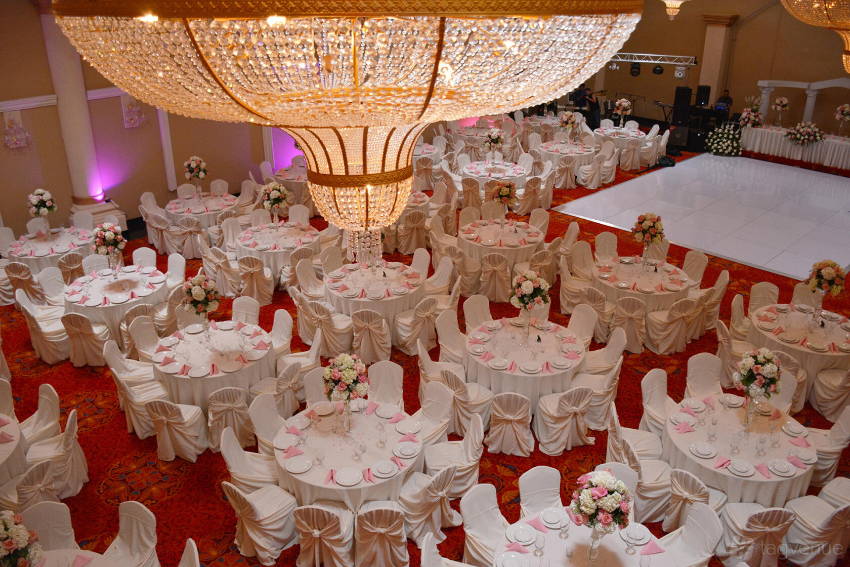 A banquet hall with round tables draped in white linens, pink accents, and a large crystal chandelier at Blue Night Banquet Hall.