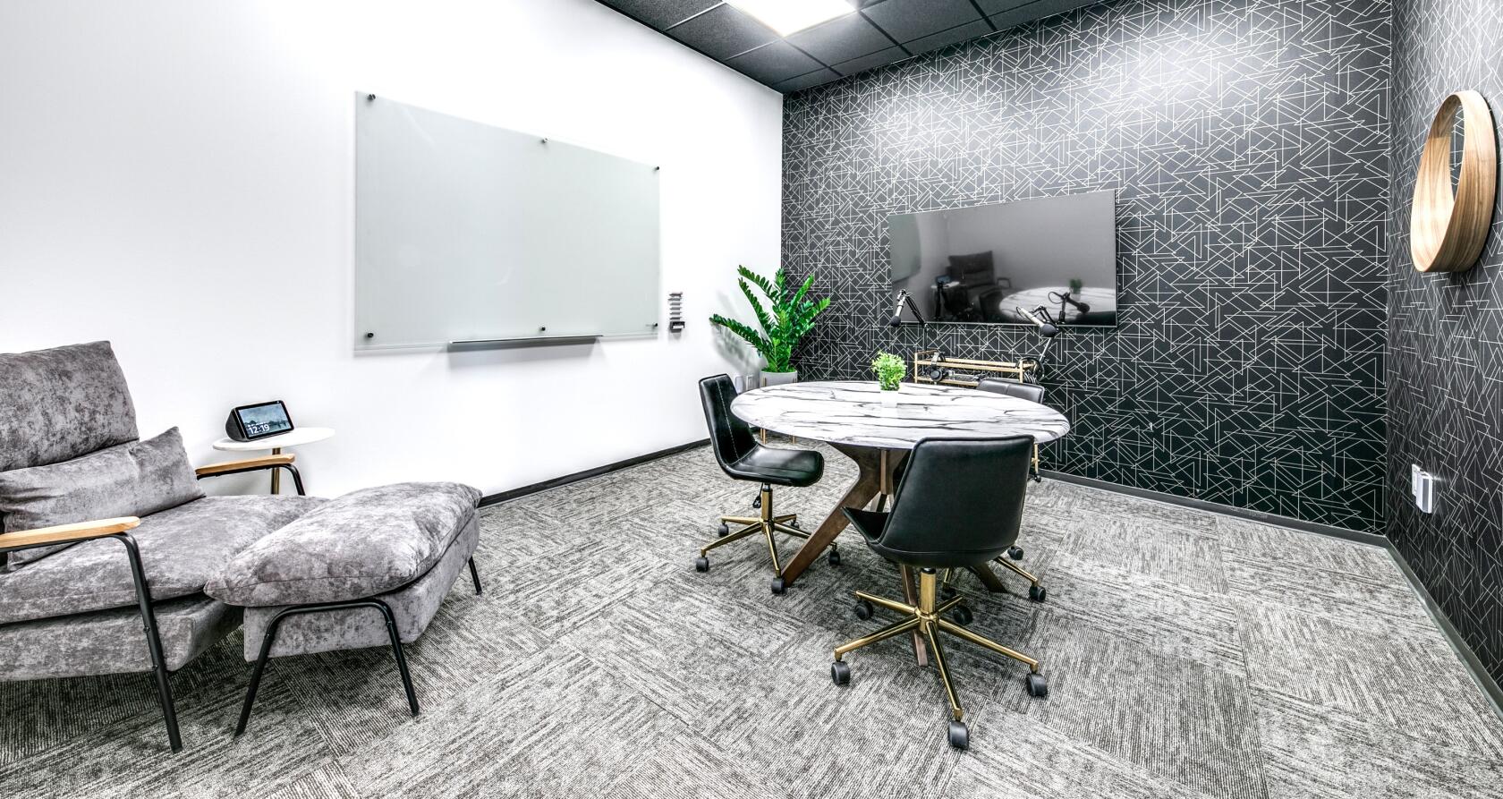 A coworking meeting room with a round marble table, wall-mounted whiteboard, large monitor, and accent wall at Lucid Private Offices - McKinney/Craig Ranch.