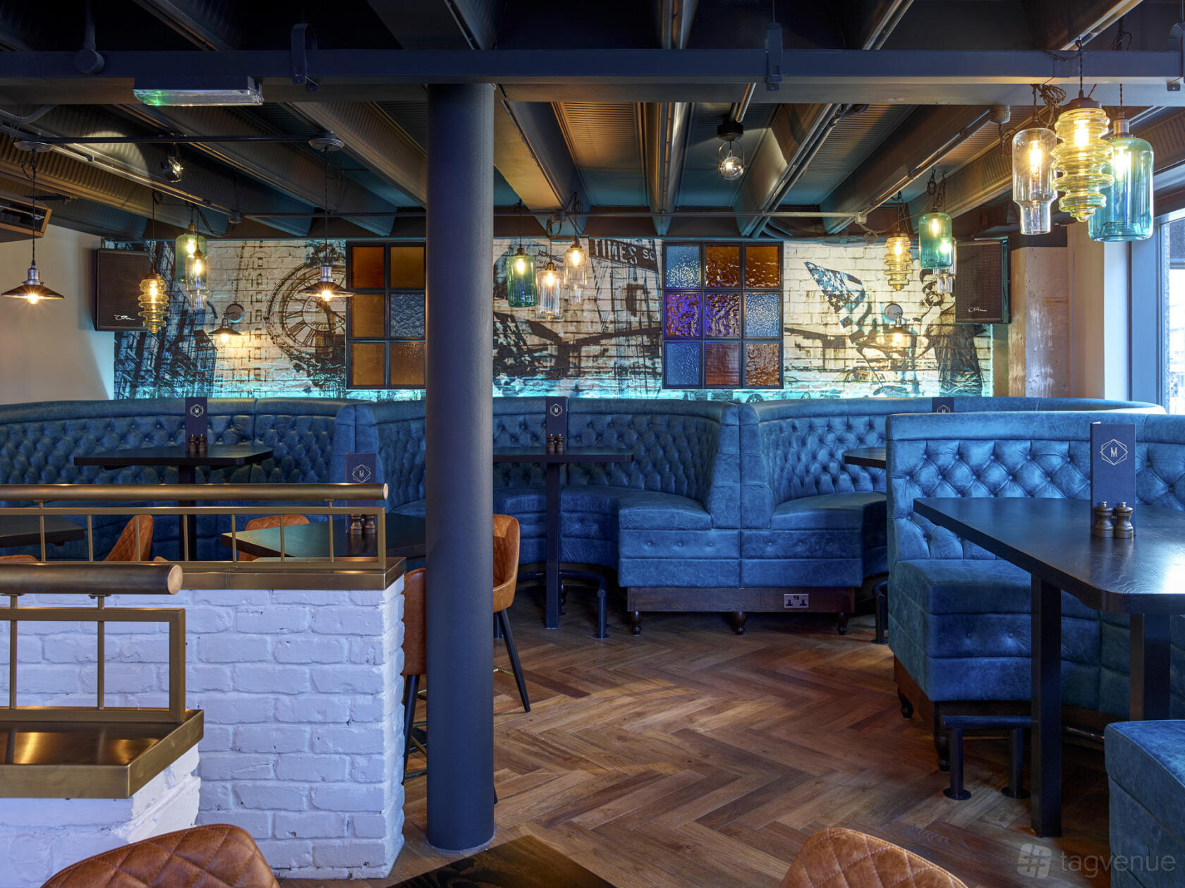 A cocktail bar with blue upholstered booth seating, pendant lighting, and a mural wall at Manahatta Greek Street.