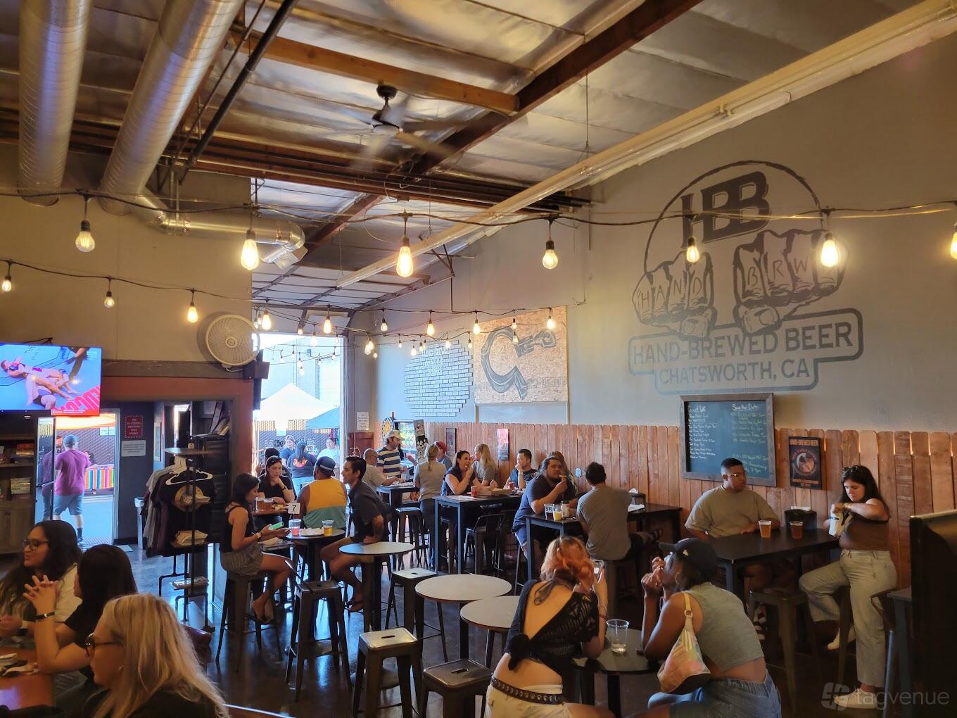 A brewery taproom with exposed ceiling beams, hanging bulbs, communal tables, and a large logo mural at Hand-Brewed Beer.