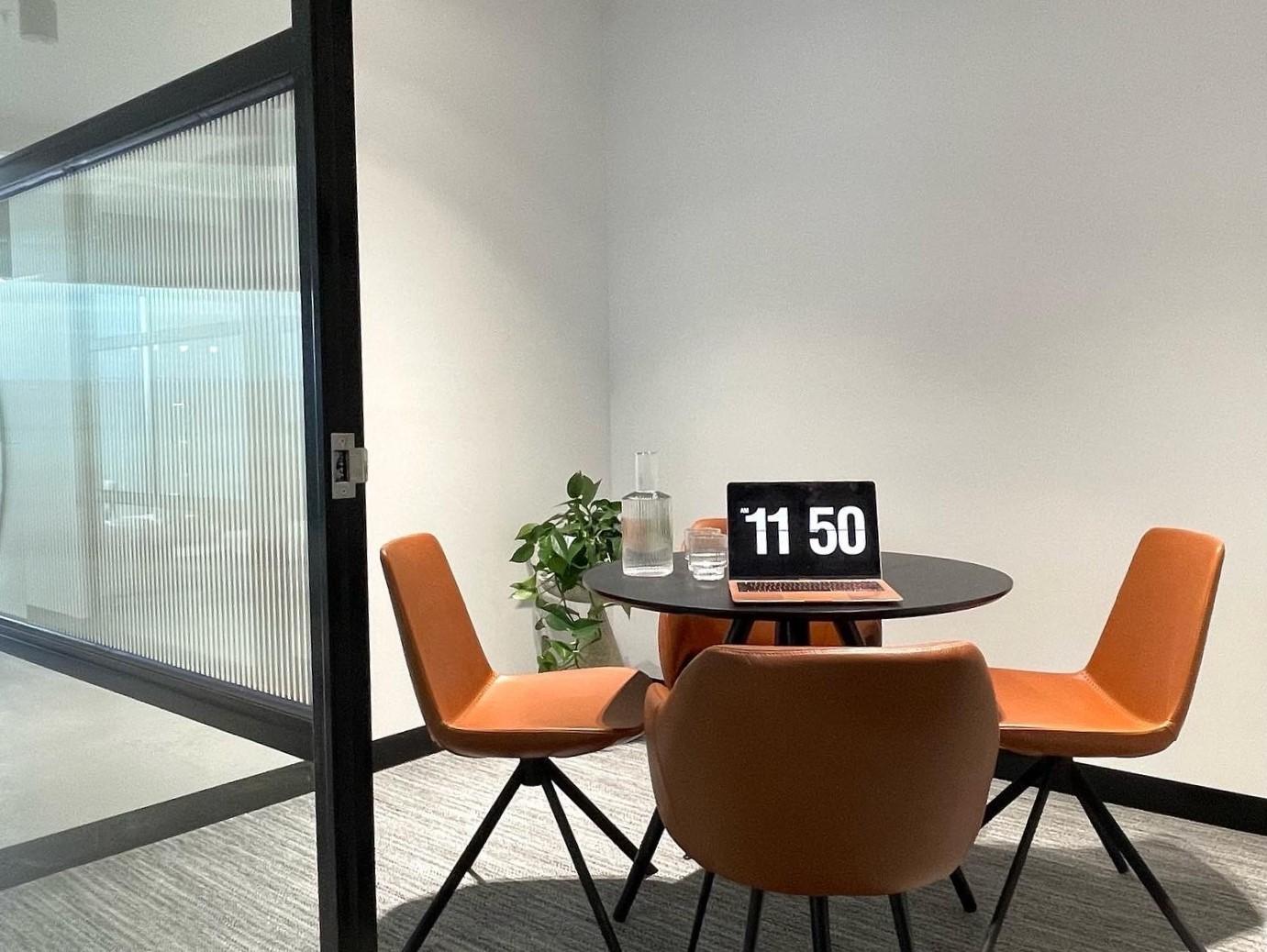 A meeting room with a round table, three tan chairs, a glass water carafe, and a potted plant at Hexa Space Coworking.