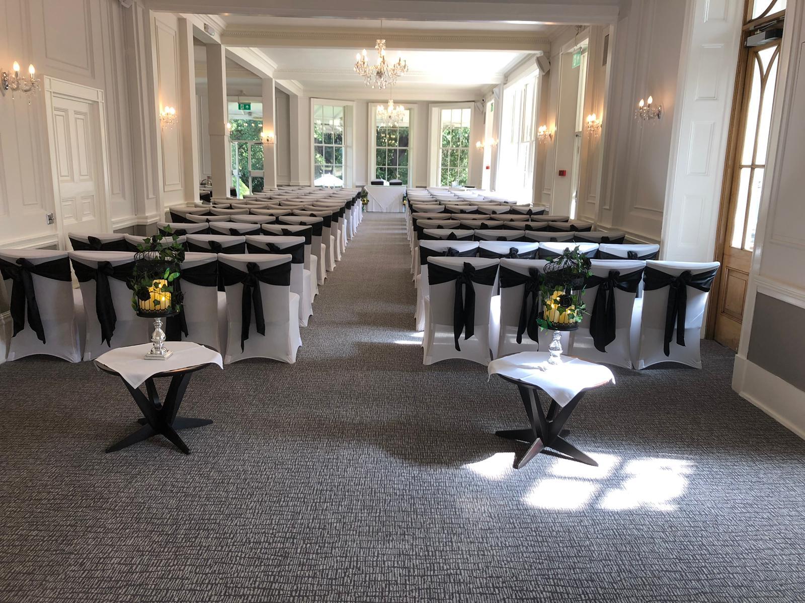 An event space with rows of white chairs adorned with black sashes, chandeliers, and tall windows at Mercure Thurrock Stifford Hall Hotel.