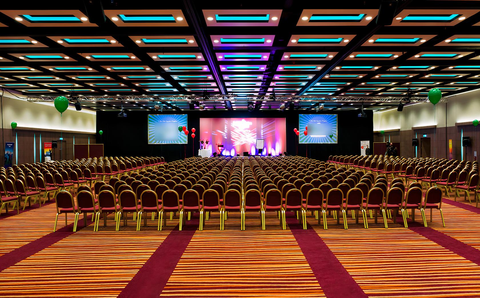 An event space in a hotel with rows of banquet chairs, colorful stage lighting, and striped carpeting at Mercure London Earl's Court.