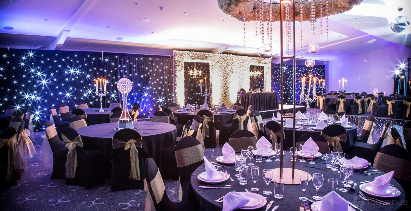 An event venue with round tables draped in black linens, starry backdrop lighting, and a white flower arch at The Hive London.