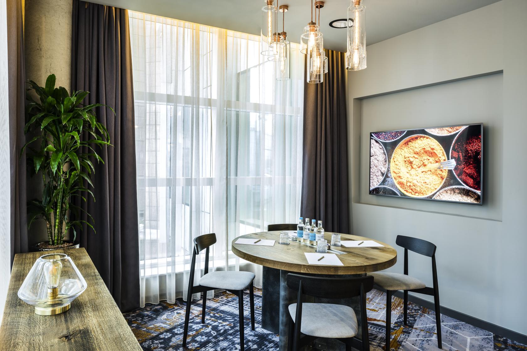 A meeting room with a round wooden table, modern pendant lights, floor-to-ceiling windows, and a wall-mounted screen at Novotel Canary Wharf.