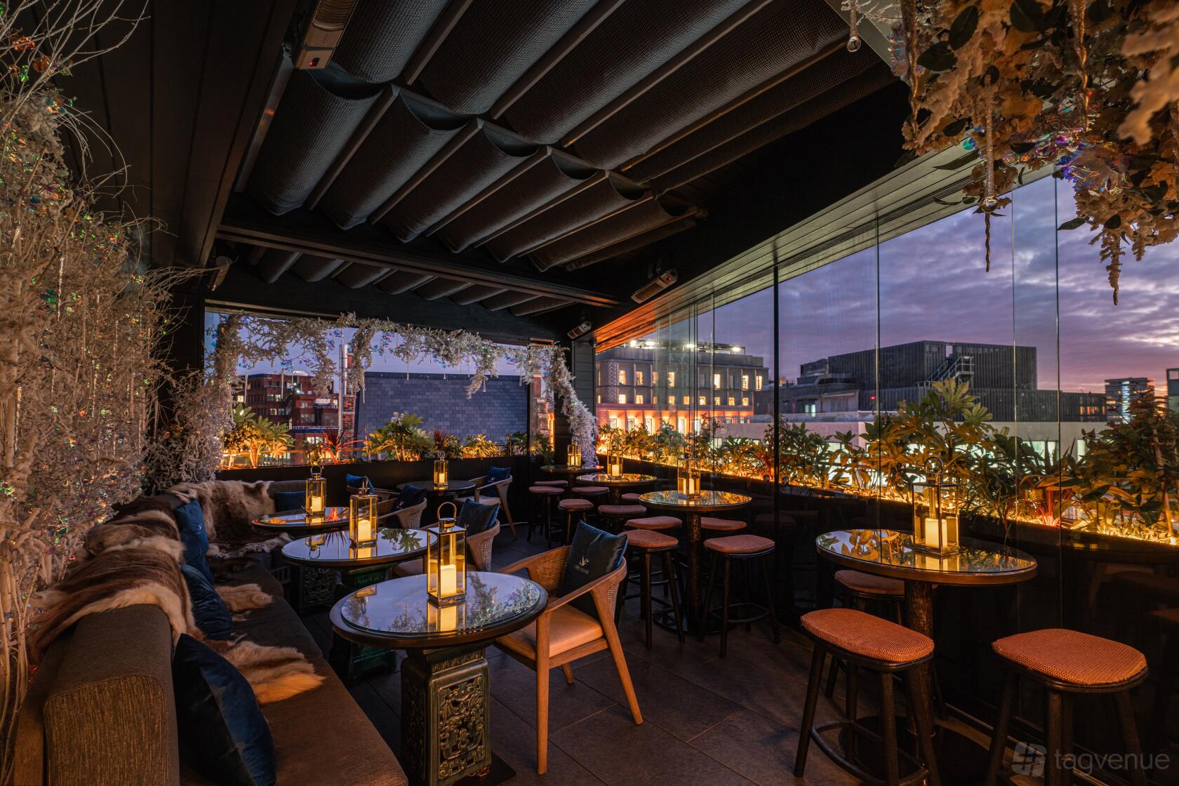 A rooftop terrace with glass walls, hanging floral arrangements, fur-accented seating, and lantern-lit tables at TATTU London.
