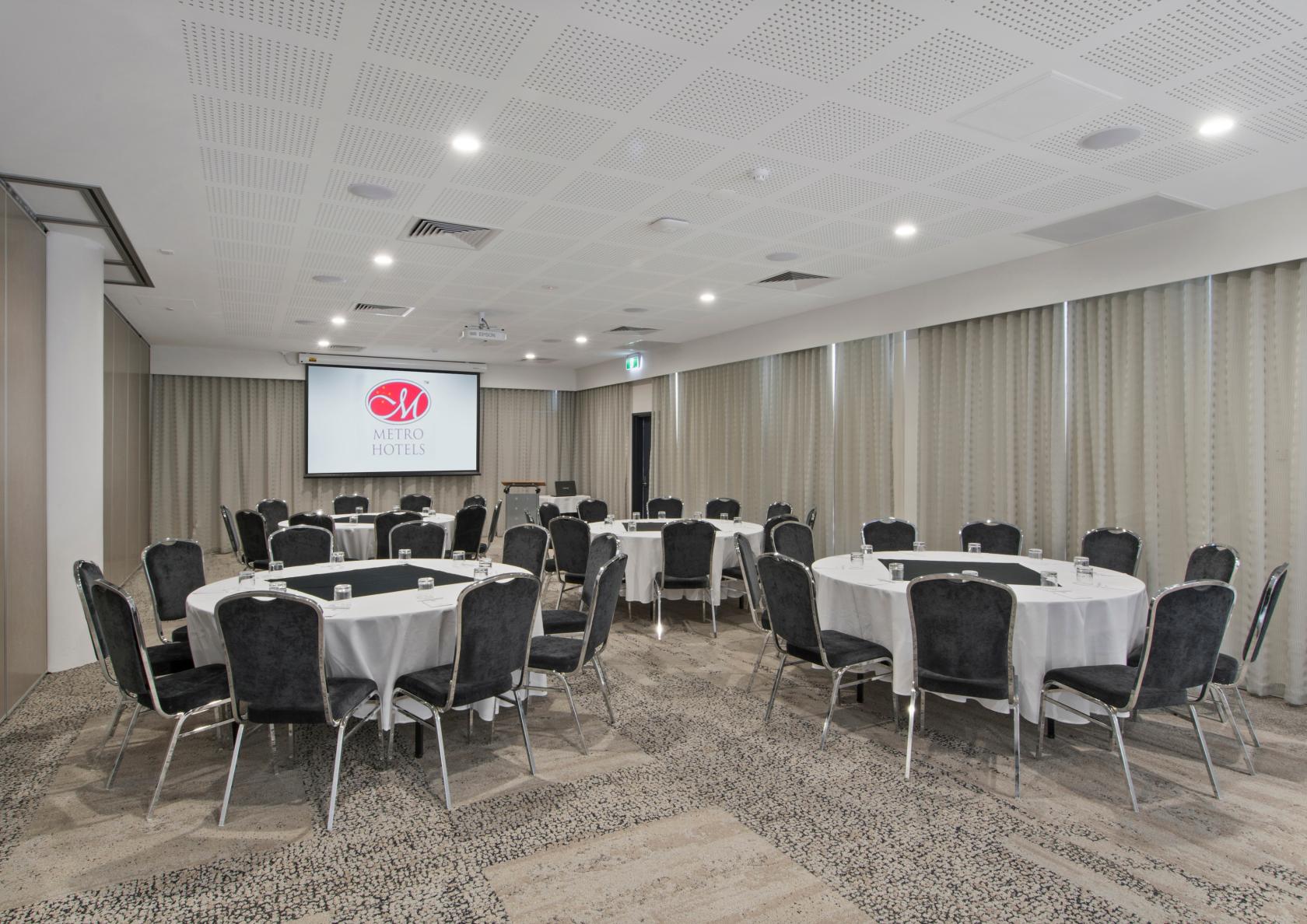 An event space with round tables, black velvet chairs, and a projection screen at Metro Hotel South Perth.