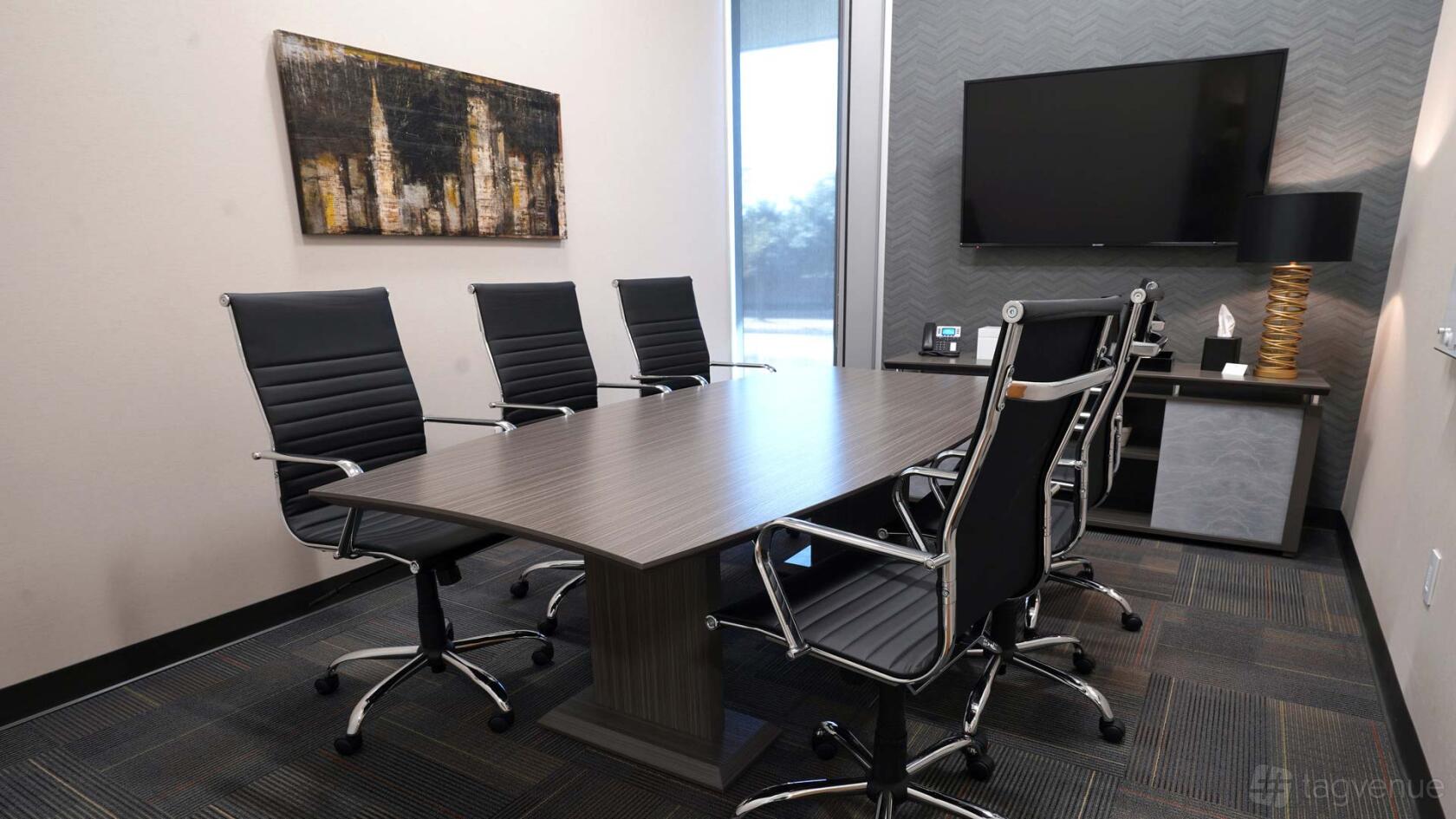 A boardroom with six black leather chairs, a wood table, wall art, and a large monitor at Executive Workspace Frisco Station.