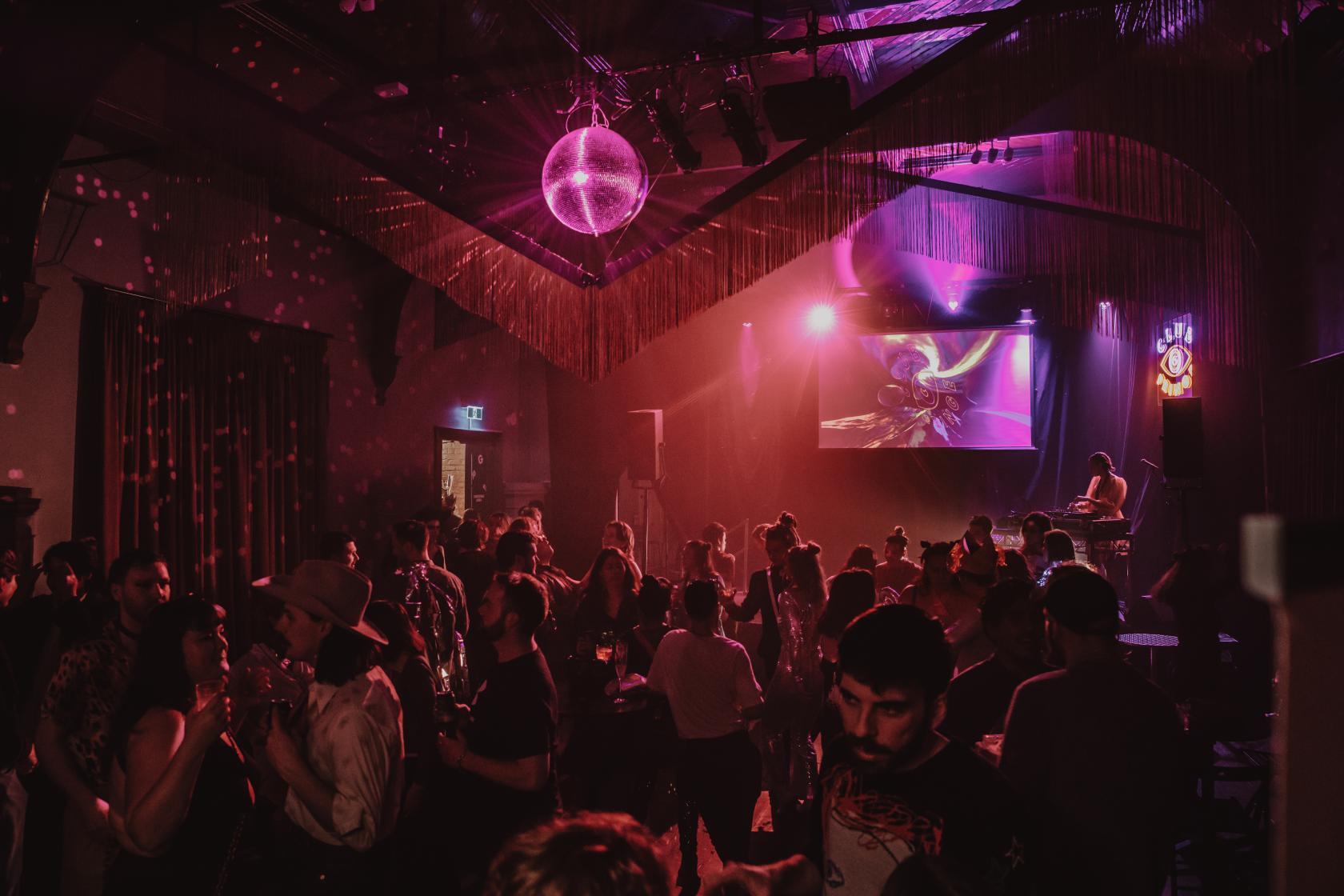 A heritage ballroom with a disco ball, stage lighting, and a large crowd dancing at Fringe Common Rooms.