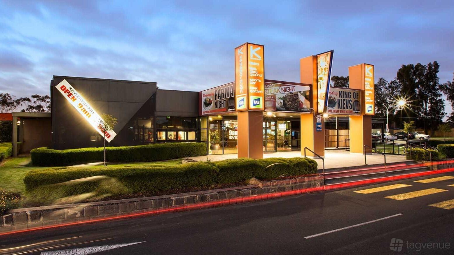 A pub exterior with illuminated signs, glass entry doors, and landscaped bushes at Keysborough Hotel.