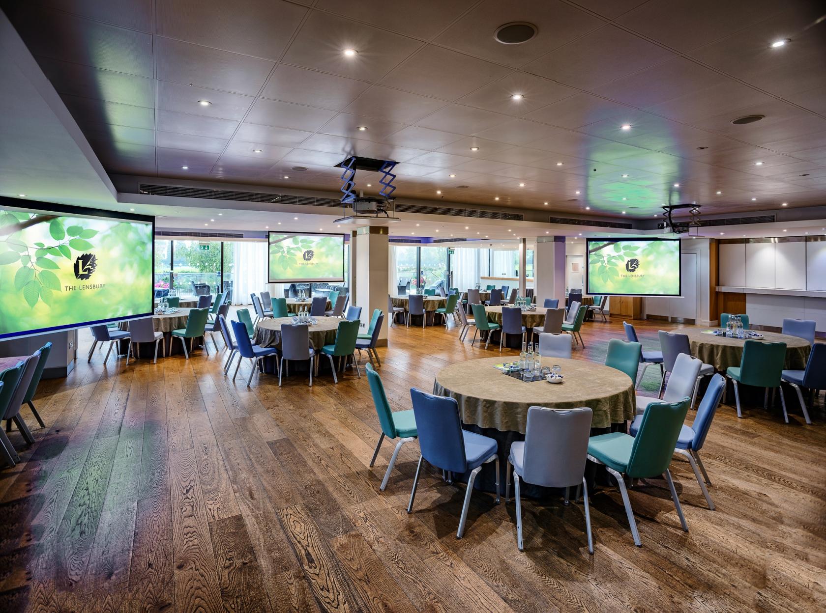 An event space with floor-to-ceiling windows, round tables, and dual projection screens at The Lensbury.