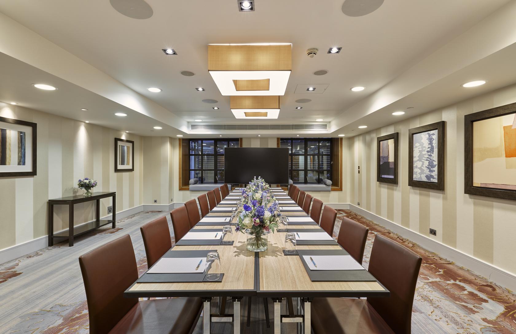 A hotel meeting room with a long conference table, brown leather chairs, and abstract art at Meetings & Events Venues.