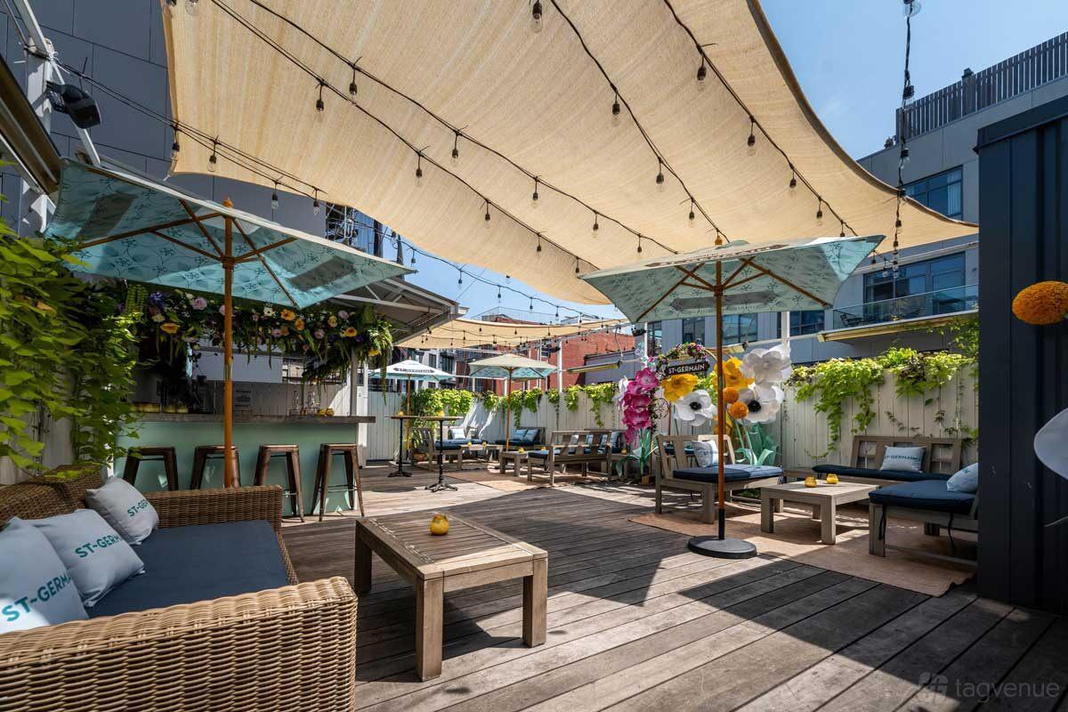 An outdoor restaurant patio with wood decking, shaded pergolas, wicker seating, and umbrella-covered tables at Ainslie.