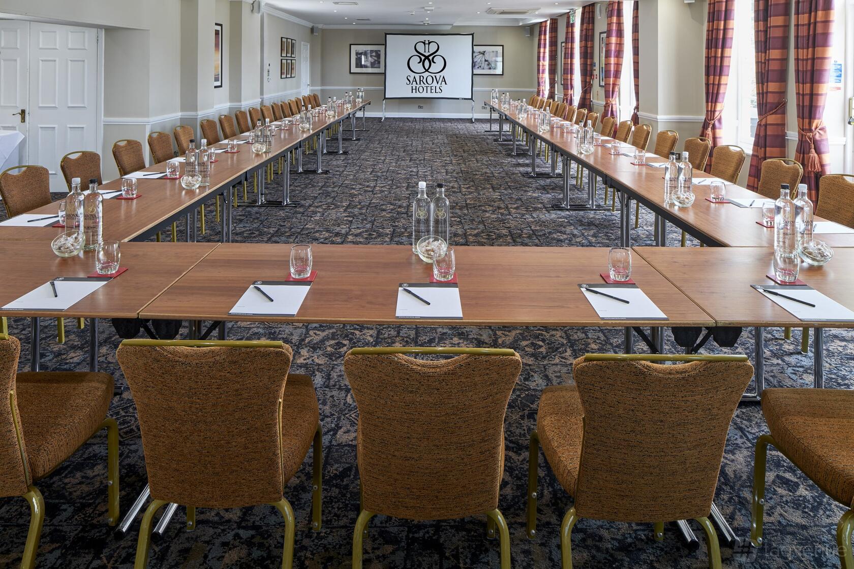 An event space with U-shaped tables, gold chairs, water bottles, and floor-to-ceiling windows at Sir Christopher Wren Hotel.