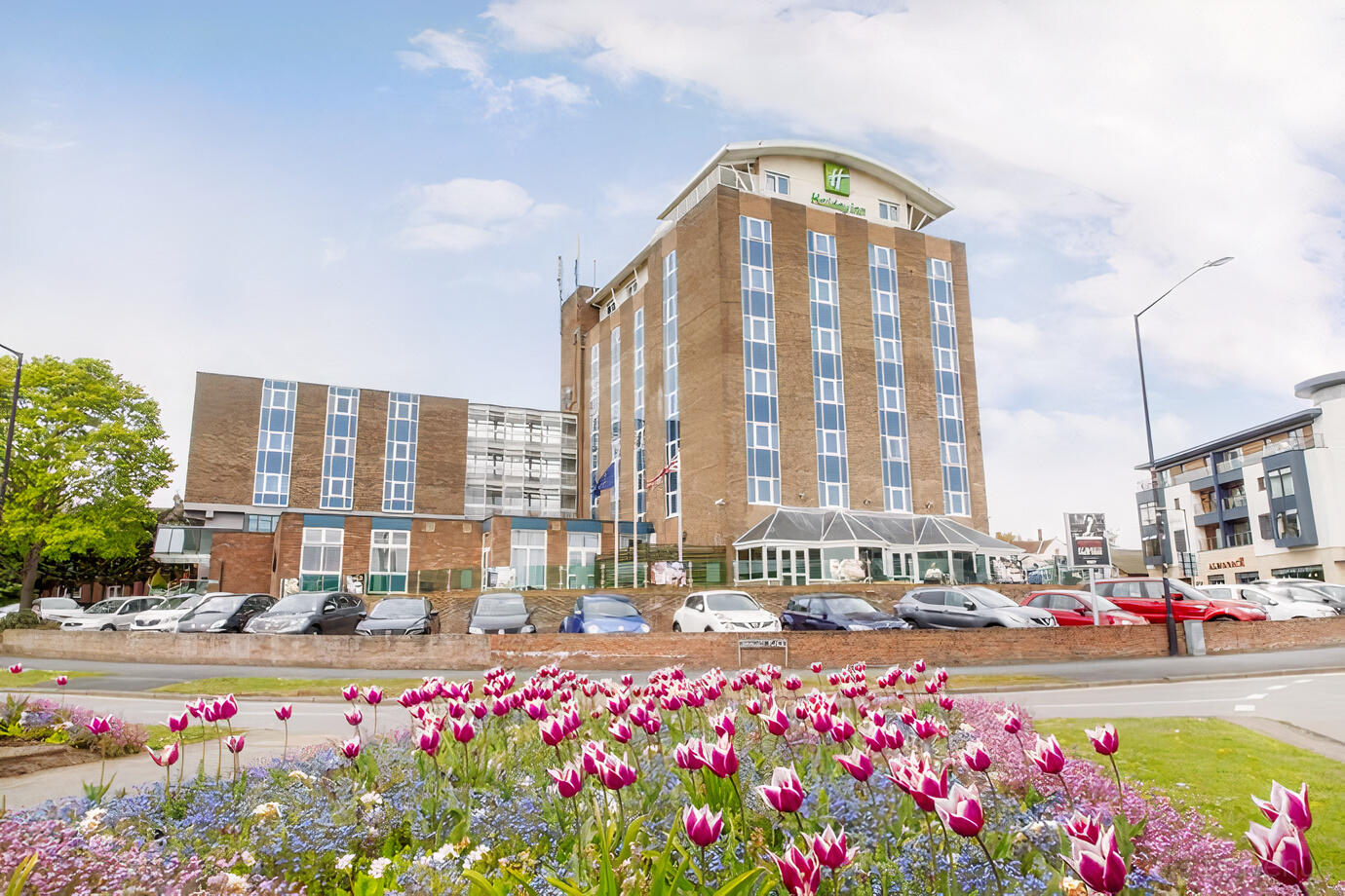 A versatile event space with a tall brick hotel exterior, glass entrance, and flower garden at Holiday Inn Kenilworth-Warwick.