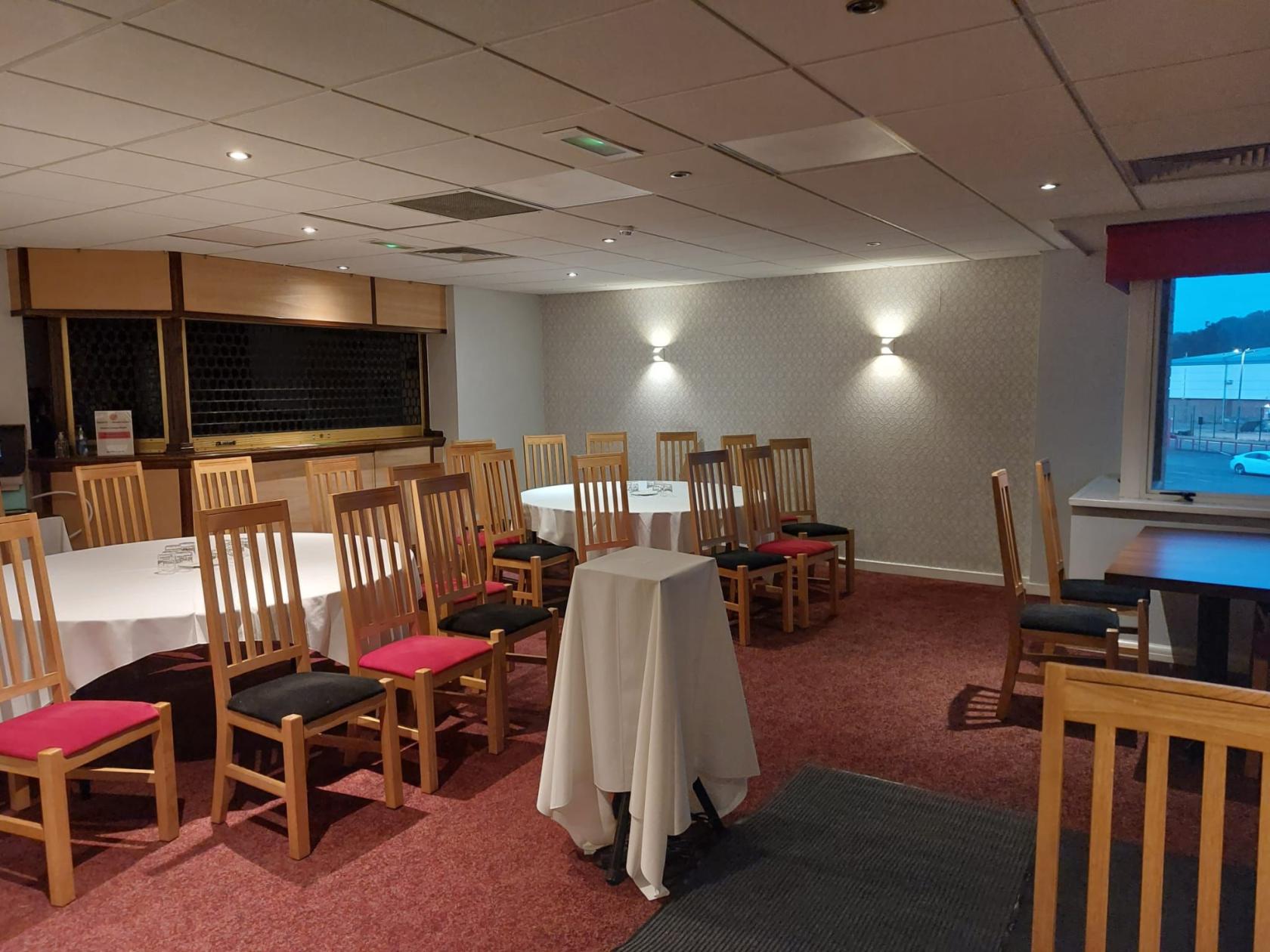 A lounge at Walsall Football Club with round tables covered in white linens, wooden chairs, and wall sconces.