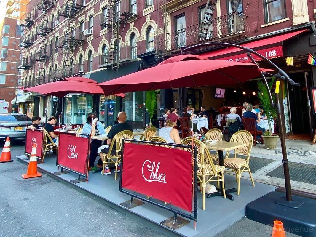 A restaurant patio with red umbrellas, rattan chairs, and branded barriers at Chelsea Ristorante Italiano.