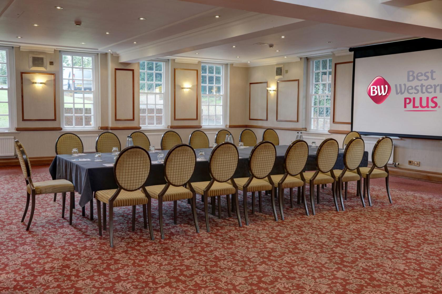 A hotel function room with large windows, patterned carpet, and a U-shaped table setup at Best Western Plus Manor Hotel.