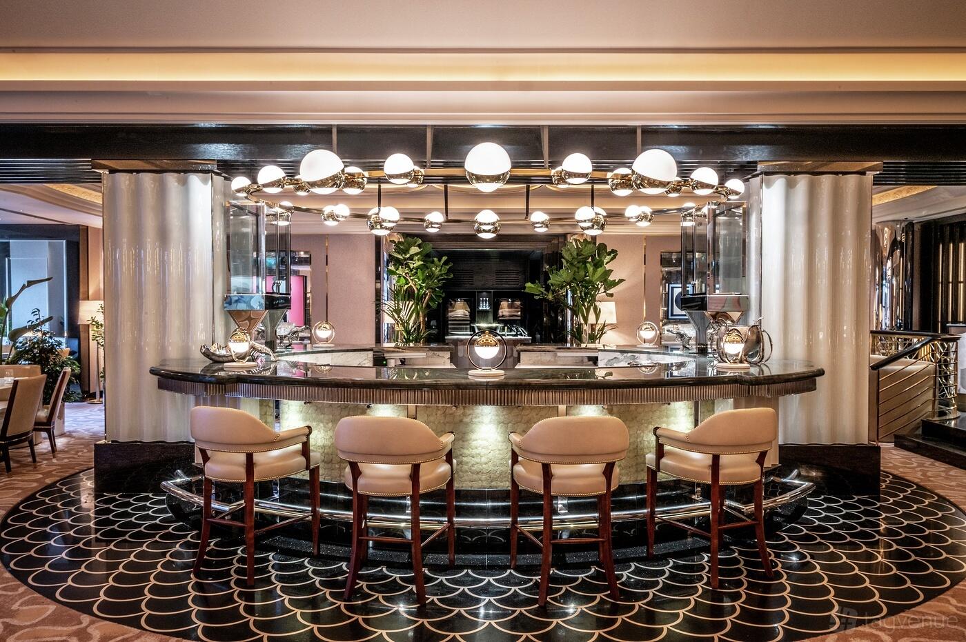 A restaurant with a central marble-topped bar, globe pendant lighting, and upholstered stools at The River Restaurant by Gordon Ramsay.