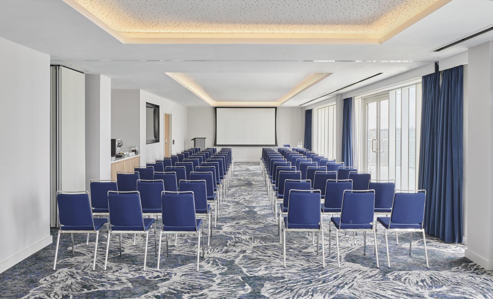 An event space with rows of blue chairs, patterned carpet, and floor-to-ceiling windows at Hyatt Regency London Stratford.