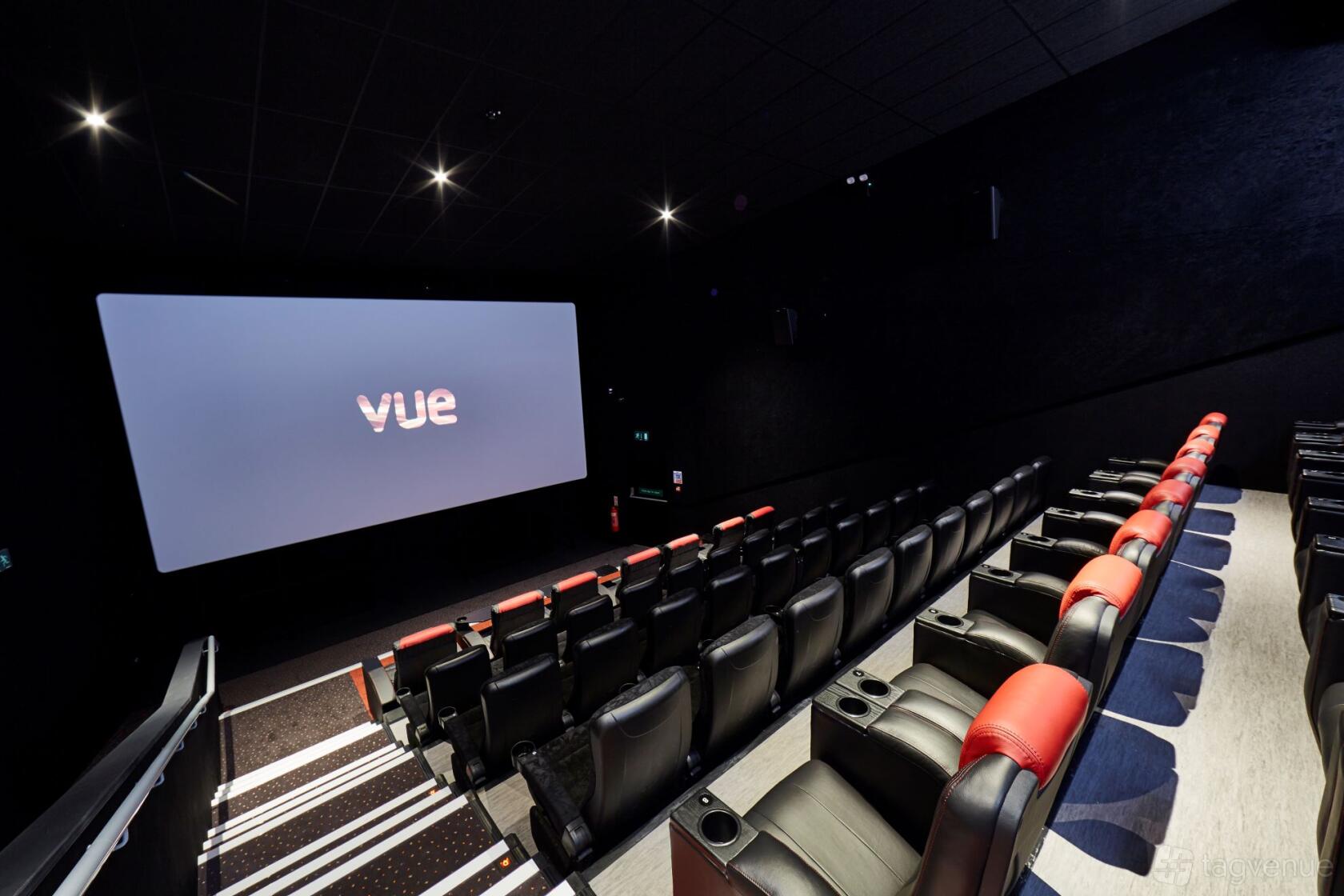 A screening room with tiered black leather seats, red accents, and a large projection screen at Vue Bedford.