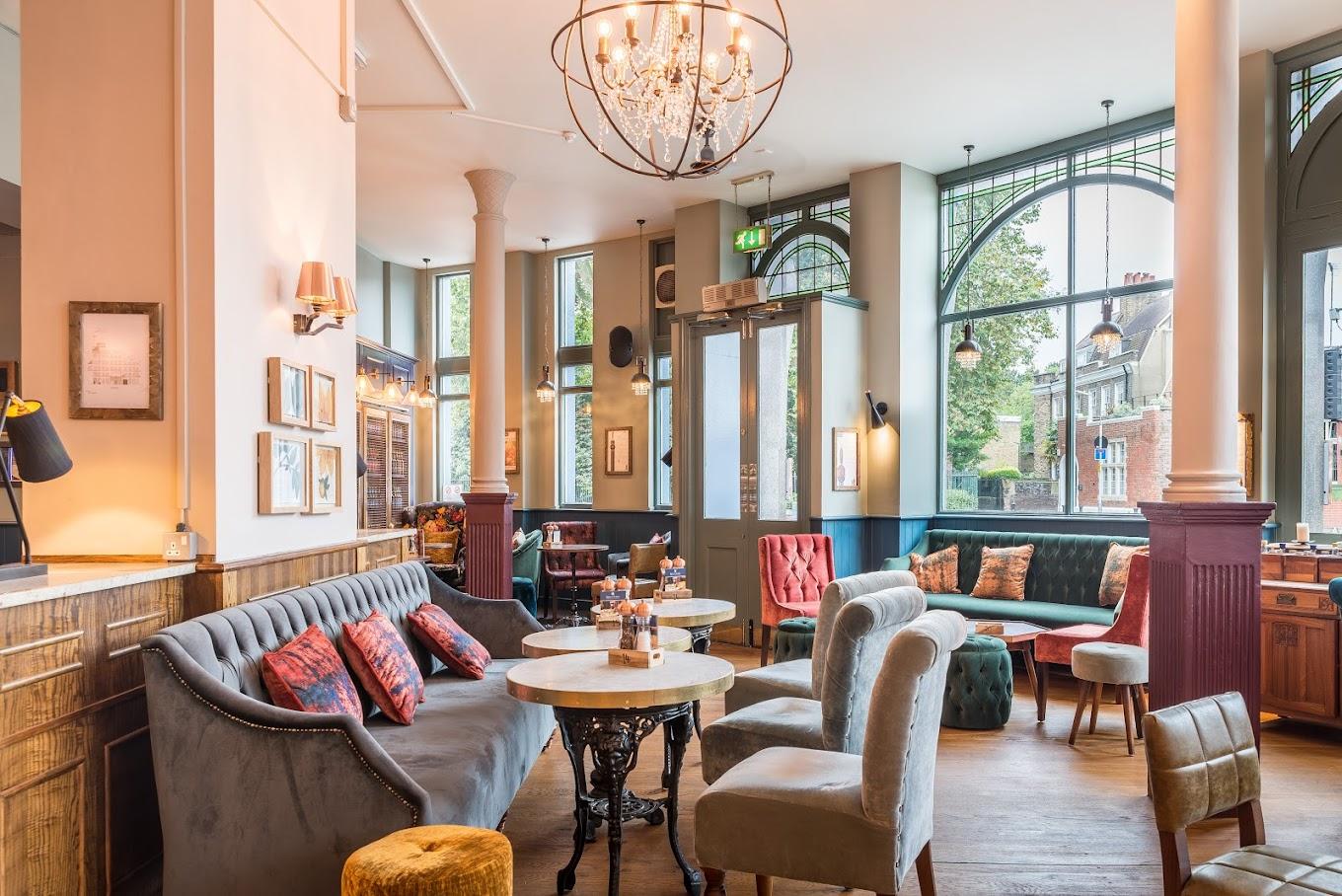A pub lounge with large arched windows, velvet sofas, round tables, and a chandelier at Kings Arms Pub.