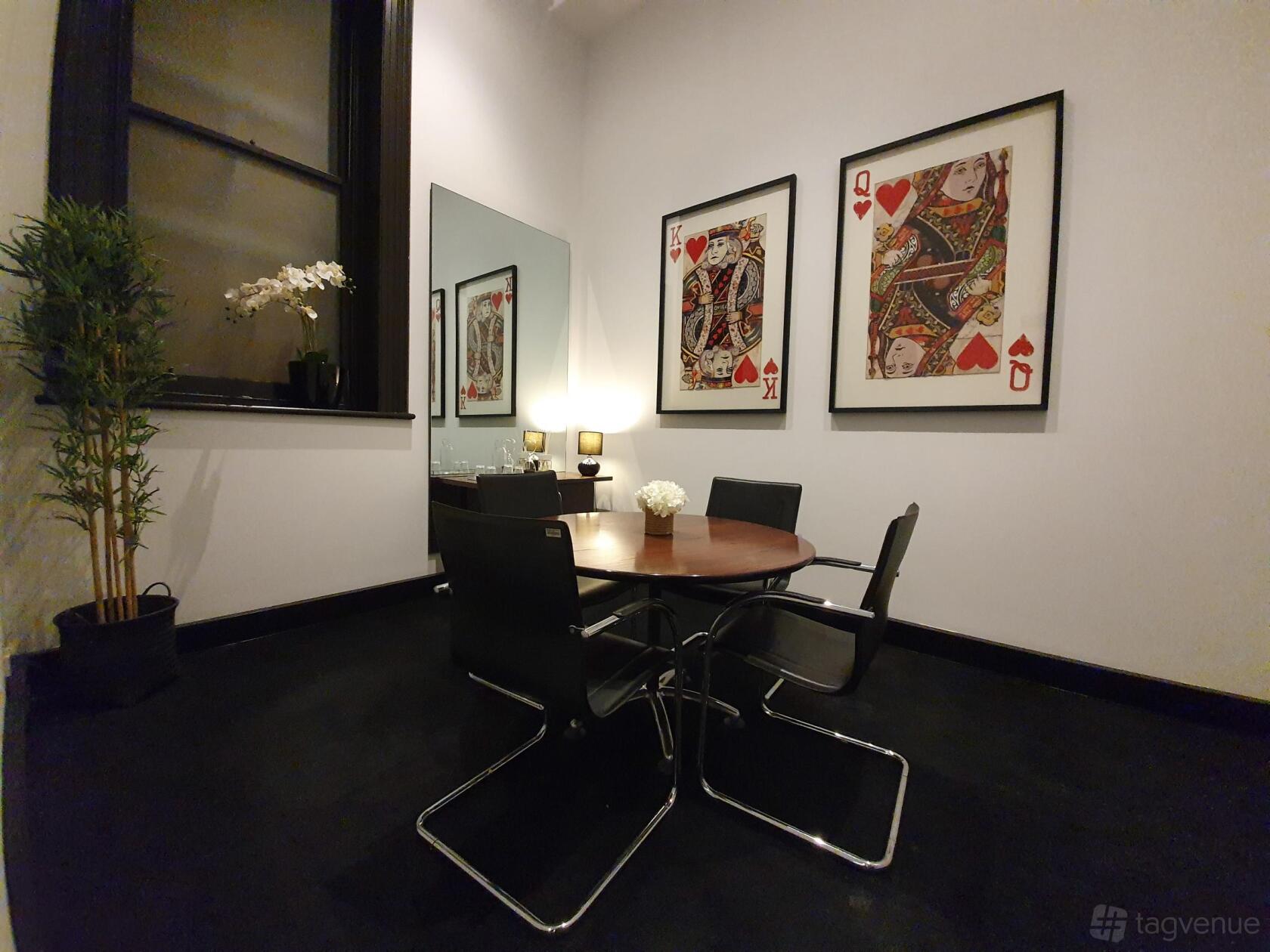A meeting room with a round table, four black chairs, framed playing card art, and potted plants at @workspaces Normanby Chambers.