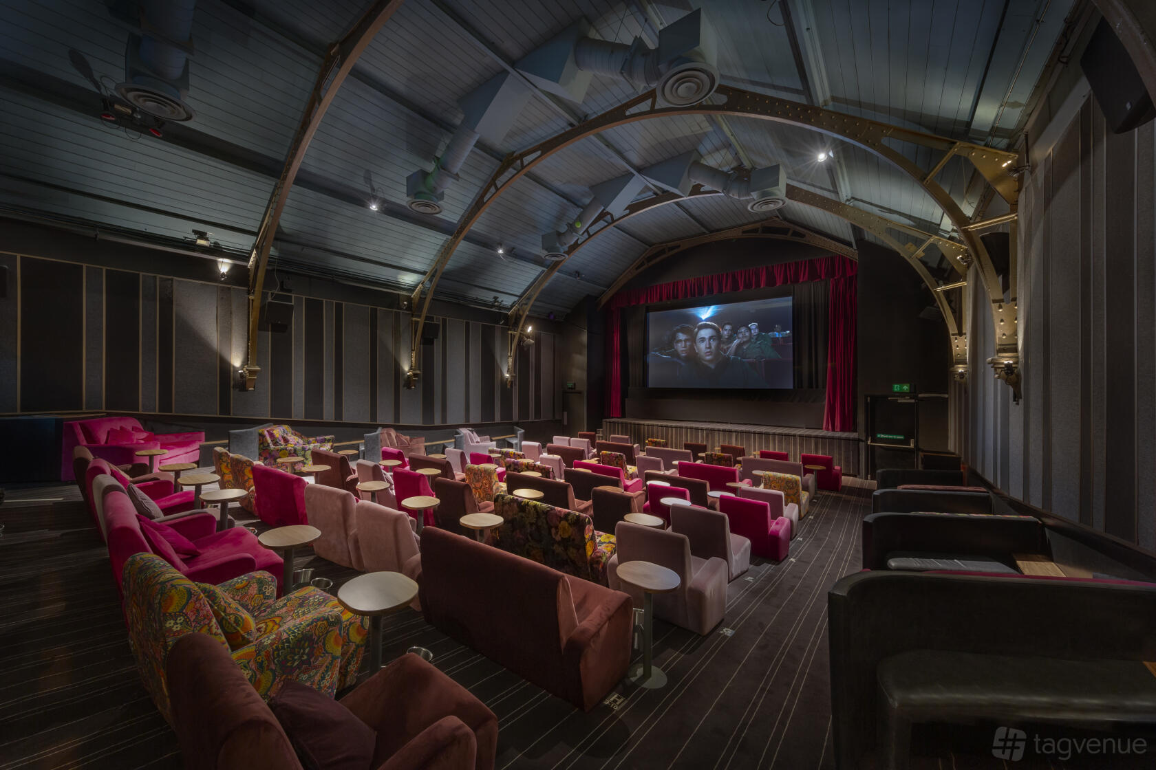 An auditorium with plush, colorful armchairs, side tables, and a large screen under arched beams at Everyman Hampstead.