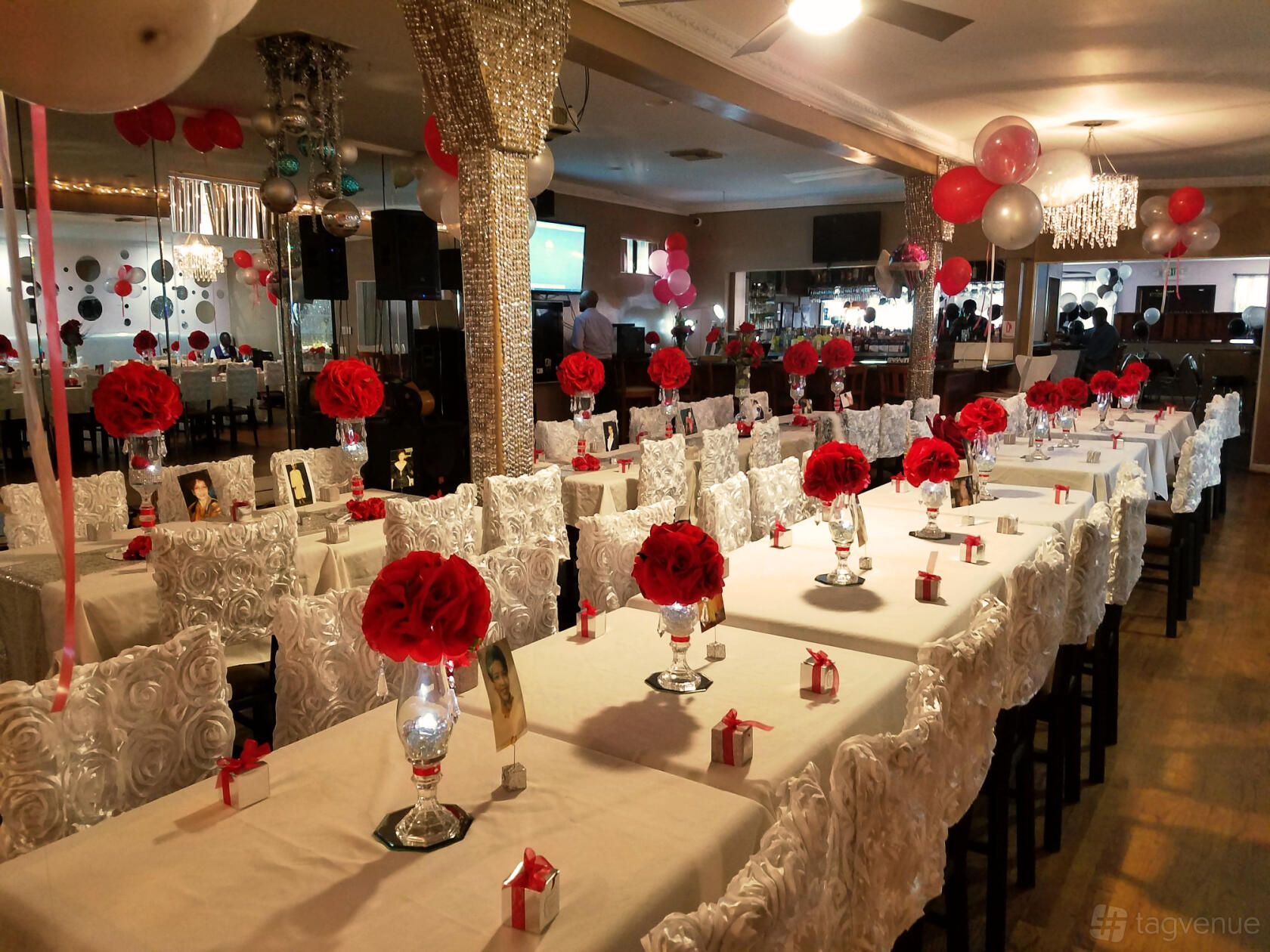 An indoor bar with long banquet tables, white chair covers, red rose centerpieces, and balloon decorations at Lavender Blue Sports Lounge.